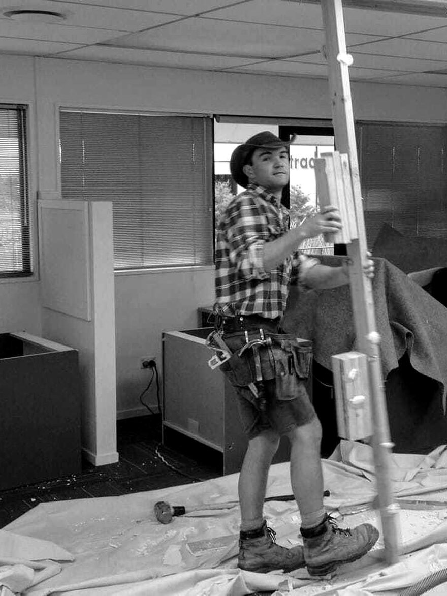 Matthew Horncastle as a young builder, in a tool belt and work boots, framing a timber partition during an office fit-out